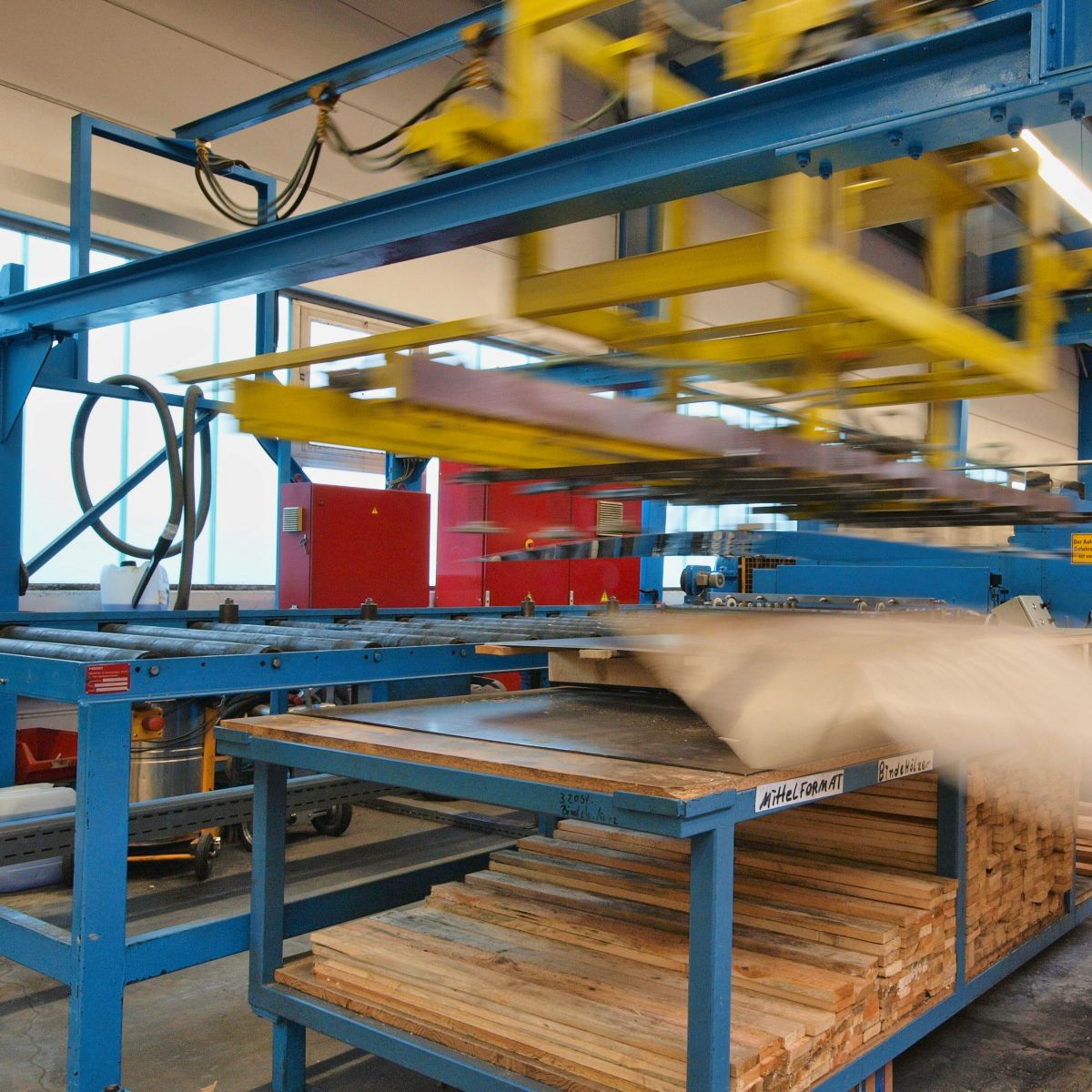 Das Bild zeigt eine Industriehalle mit einer Maschine aus einer blauen Stahlkonstruktion mit einer Rollenbahn. Daneben ein Regal mit zwei Ebenen. Unten sind Holzprofile gelagert, oben liegt ein Stapel von Flachblechen auf Holzprofilen flach auf. Ein gelber Schlitten an der Stahlkonstruktion bewegt sich im Bild, was durch Unschärfe sichtbar ist. Er hat mit Saugnäpfen ein Blech gepackt, um es auf die Rollenbahn zu heben. Nach rechts fällt ein Blatt Schutzpapier weg, das zwischen den Blechen liegt.