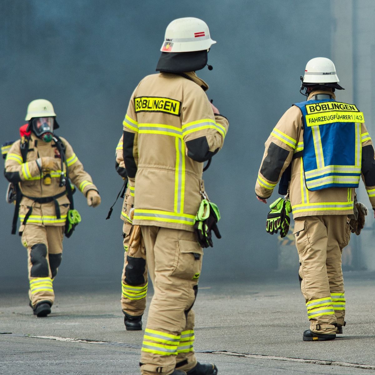 Das Bild zeigt vier Feuerwehrleute der Feuerwehr Böblingen in beigen Uniformen mit gelb-silbernen Reflektorstreifen und weißen Helmen. Zwei laufen auf den Fotografen zu, sie haben Pressluftatmer über dem Gesicht und die Luftflaschen auf dem Rücken. Hinter ihnen ist in dichtem Rauch schemenhaft die Öffnung einer Werkshalle zu erkennen. Zwei im Vordergrund stehen mit dem Rücken zum Betrachter und erwarten den Atemschutz-Trupp. Einer der beiden trägt zusätzlich eine blau-gelbe Warnweste, auf der zu lesen ist „Fahrzeugführer 1/43“.