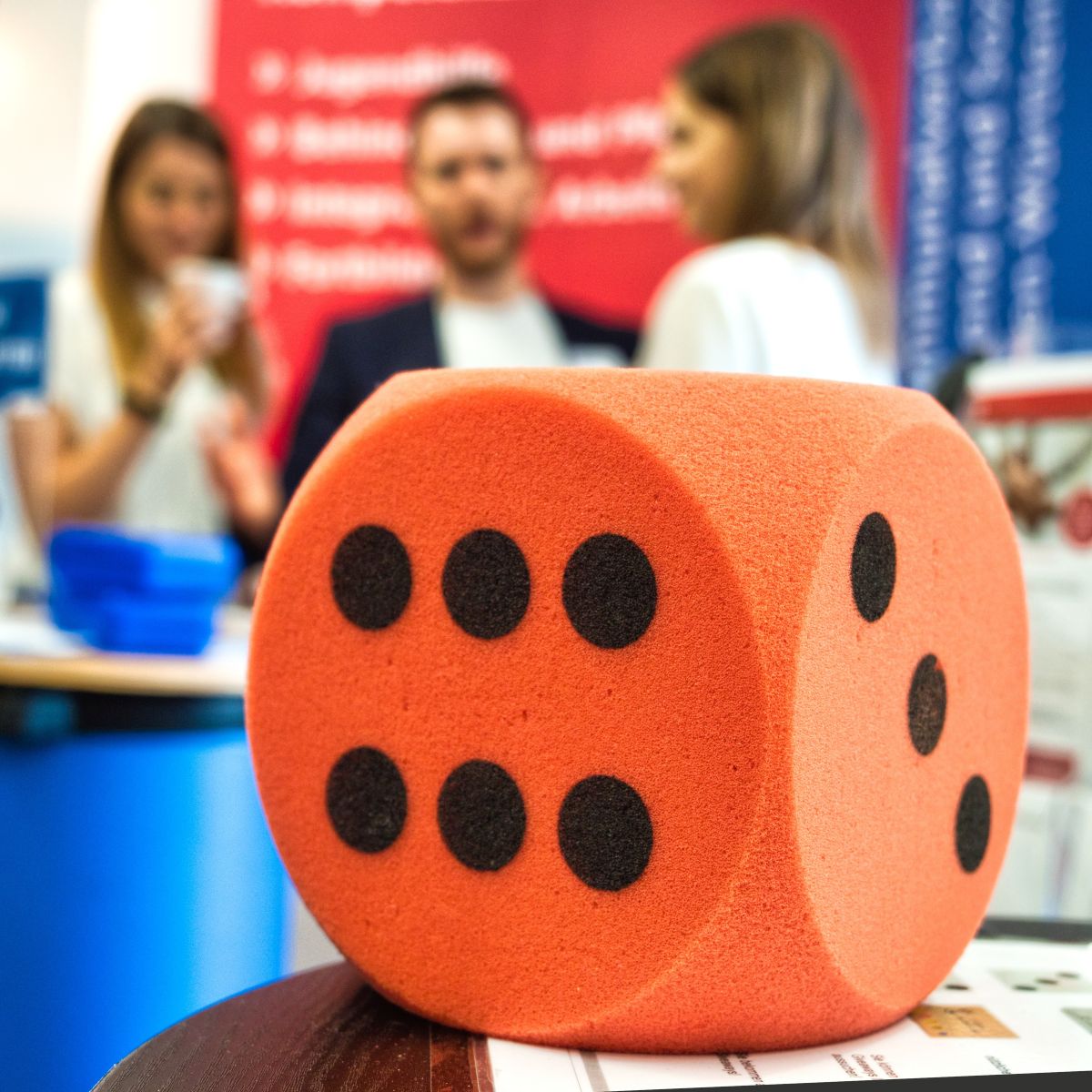 Das Bild zeigt groß im Vordergrund einen roten Schaumstoffwürfel auf einem Holztisch, auf dem die Seiten mit sechs und mit drei Augen zu sehen sind. Unscharf im Hintergrund stehen zwei Frauen und ein Mann an einem Messestand.